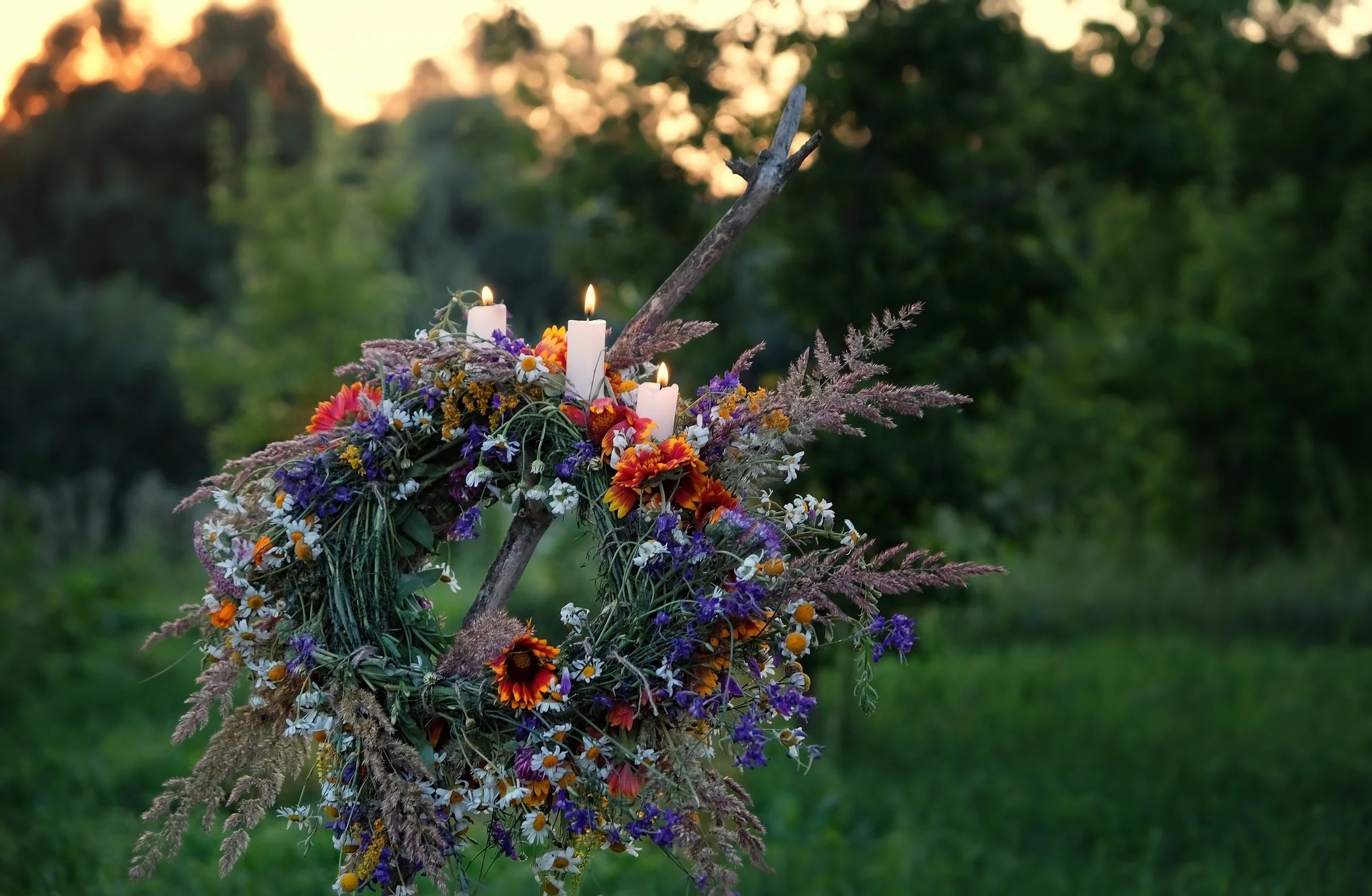 A wildflower wreath with lit candles displayed on a wooden fork outdoors at sunset — perfect Beltane home ideas for honouring the fire festival with natural florals and candlelight